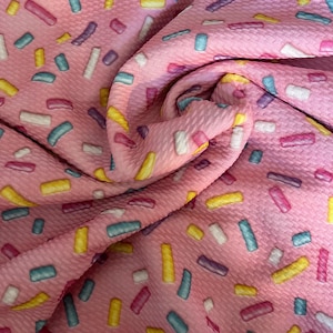 May include: Pink fabric with a sprinkle pattern. The sprinkles are in various colors, including yellow, blue, pink, and white.