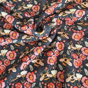 May include: Close-up of a dark fabric with a repeating pattern of bees, flowers, and foliage. The flowers are in shades of pink, red, and white, while the bees have yellow and black stripes. The fabric has a textured appearance.