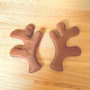 Könnte beinhalten: Zwei braune Rentiergeweihe aus Filz mit einer aufgenähten Kontur, die auf einer hellen Holzoberfläche liegen. Die Geweihe sind symmetrisch und haben ein strukturiertes Aussehen, was auf ein weiches Material hindeutet. Ideal für Bastelarbeiten oder Kostüme.