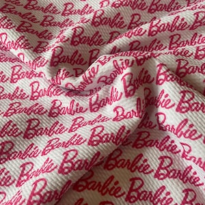 May include: White fabric with a repeating pattern of the word "Barbie" in a vibrant pink script. The fabric has a textured, ribbed appearance, with the word "Barbie" densely printed across the surface.