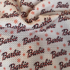 May include: White fabric with a red and black checkered pattern featuring the word "Barbie" repeated throughout. The fabric has a snowflake pattern.