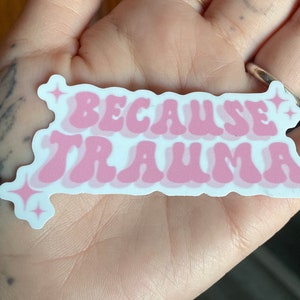 K&ouml;nnte beinhalten: Rosa-wei&szlig;er Aufkleber mit dem Text "Because Trauma" in einer Retro-Schriftart mit funkelnden Sternen auf beiden Seiten.