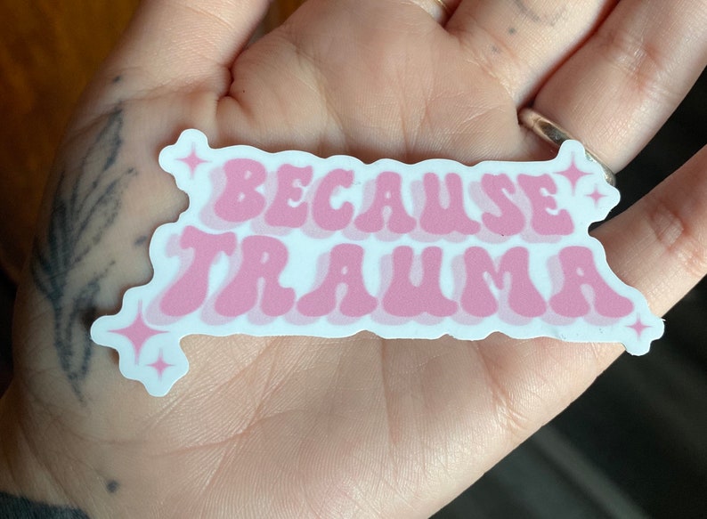 K&ouml;nnte beinhalten: Rosa-wei&szlig;er Aufkleber mit dem Text "Because Trauma" in einer Retro-Schriftart mit funkelnden Sternen auf beiden Seiten.