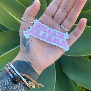 K&ouml;nnte beinhalten: Ein rosa-wei&szlig;er Aufkleber mit dem Text "Because Trauma" in einer Retro-Schriftart. Der Aufkleber wird in einer Hand mit einem silbernen Ring am Zeigefinger gehalten.
