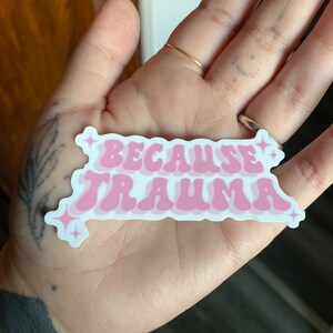 K&ouml;nnte beinhalten: Rosa-wei&szlig;er Aufkleber mit dem Text "Because Trauma" in einer Retro-Schriftart. Der Aufkleber hat eine gl&auml;nzende Oberfl&auml;che und zwei kleine Glitzerakzente.