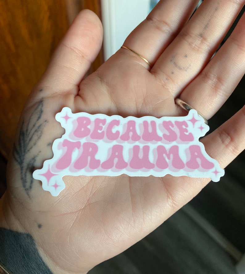 K&ouml;nnte beinhalten: Rosa-wei&szlig;er Aufkleber mit dem Text "Because Trauma" in einer Retro-Schriftart. Der Aufkleber hat eine gl&auml;nzende Oberfl&auml;che und zwei kleine Glitzerakzente.