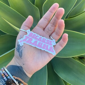 K&ouml;nnte beinhalten: Rosafarbener Aufkleber mit dem Text "Because Trauma" in einer Retro-Schriftart mit Sternen auf beiden Seiten. Der Aufkleber wird in einer Hand mit einem Tattoo am Handgelenk gehalten.