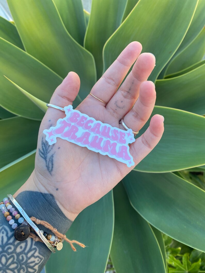 K&ouml;nnte beinhalten: Rosafarbener Aufkleber mit dem Text "Because Trauma" in einer Retro-Schriftart mit Sternen auf beiden Seiten. Der Aufkleber wird in einer Hand mit einem Tattoo am Handgelenk gehalten.
