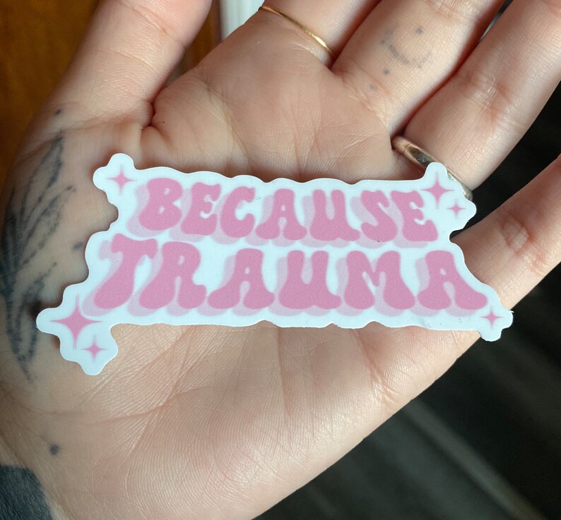 K&ouml;nnte beinhalten: Rosafarbener Aufkleber mit dem Text "Because Trauma" in einer Retro-Schriftart mit wei&szlig;en Sternen auf beiden Seiten.