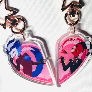 Puede incluir: Dos llaveros en forma de corazón con personajes de dibujos animados. Uno tiene un personaje azul y blanco, el otro un personaje rojo y negro. Ambos tienen fondos rosas y adornos dorados en forma de estrella.