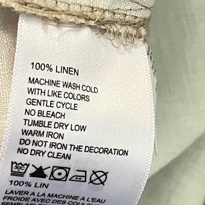 May include: A white fabric tag with black text detailing care instructions for a linen garment. The tag reads "100% LINEN" and includes washing, drying, and ironing guidelines, along with care symbols.