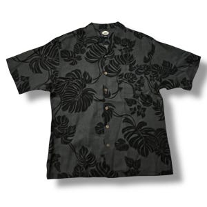 Puede incluir: Camisa gris oscuro de manga corta con un estampado floral negro. La camisa tiene cuello y botones de madera. El dise&ntilde;o floral incluye hojas grandes y follaje tropical.
