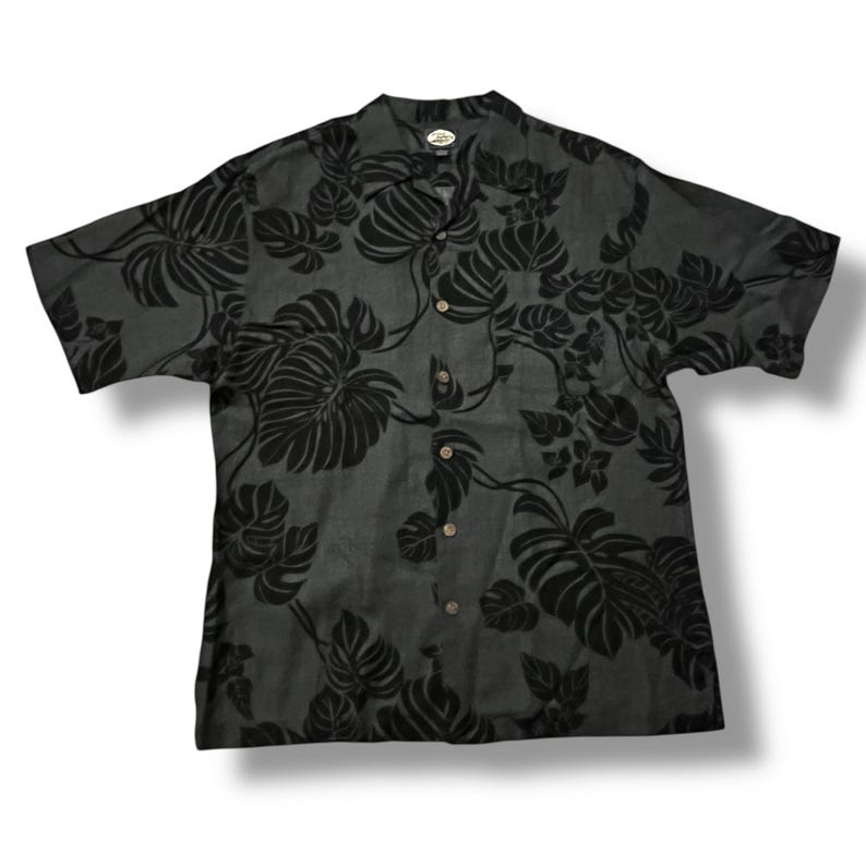 Puede incluir: Camisa gris oscuro de manga corta con un estampado floral negro. La camisa tiene cuello y botones de madera. El dise&ntilde;o floral incluye hojas grandes y follaje tropical.