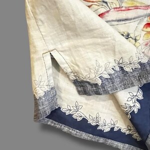 May include: Close-up of a cream-colored garment with a blue and gray border. The border is decorated with a leaf design. The garment has a split at the sleeve. The fabric appears to be linen or a similar material.