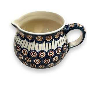 May include: A ceramic creamer pitcher featuring a rounded body and a side handle. The exterior is decorated with a repeating pattern of dark blue, white, and brown concentric circles, accented by vertical blue lines against a cream background.