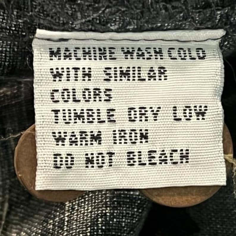 Puede incluir: Una etiqueta de tela blanca con texto negro, que incluye instrucciones de cuidado: "MACHINE WASH COLD WITH SIMILAR COLORS, TUMBLE DRY LOW, WARM IRON, DO NOT BLEACH." La etiqueta est&aacute; sujeta a una prenda con dos botones marrones.