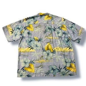 May include: A short-sleeved button-down shirt with a floral pattern. The shirt features a light gray background with large teal and yellow flowers. The shirt has a classic collar and a relaxed fit.