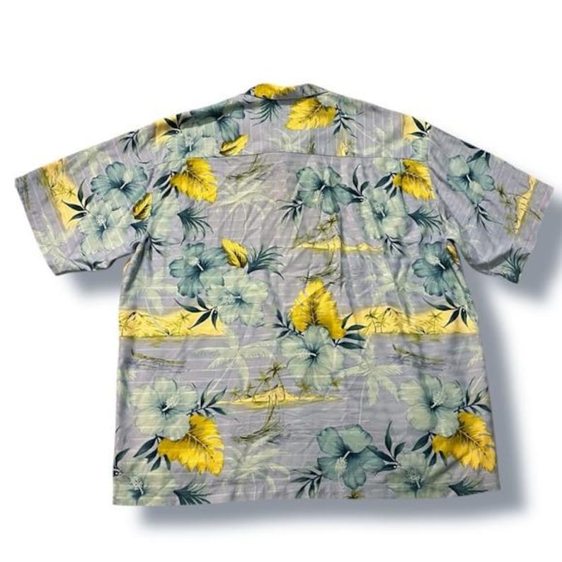 May include: A short-sleeved button-down shirt with a floral pattern. The shirt features a light gray background with large teal and yellow flowers. The shirt has a classic collar and a relaxed fit.