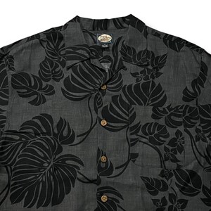 Puede incluir: Camisa gris oscuro con estampado floral negro. La camisa tiene cuello, bolsillo en el pecho y botones de madera. El dise&ntilde;o floral incluye hojas grandes y flores.