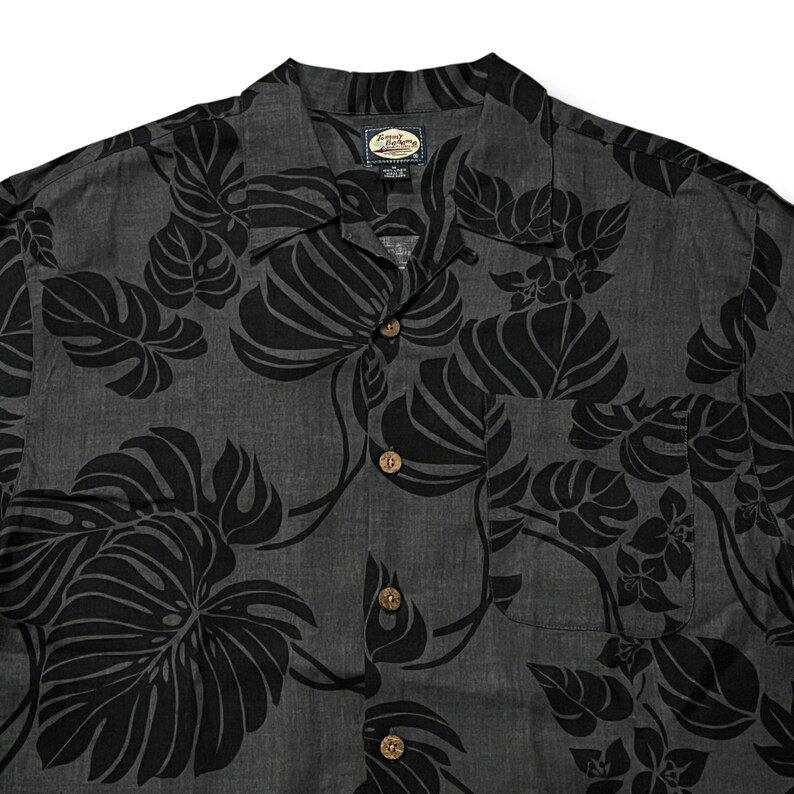 Puede incluir: Camisa gris oscuro con estampado floral negro. La camisa tiene cuello, bolsillo en el pecho y botones de madera. El dise&ntilde;o floral incluye hojas grandes y flores.