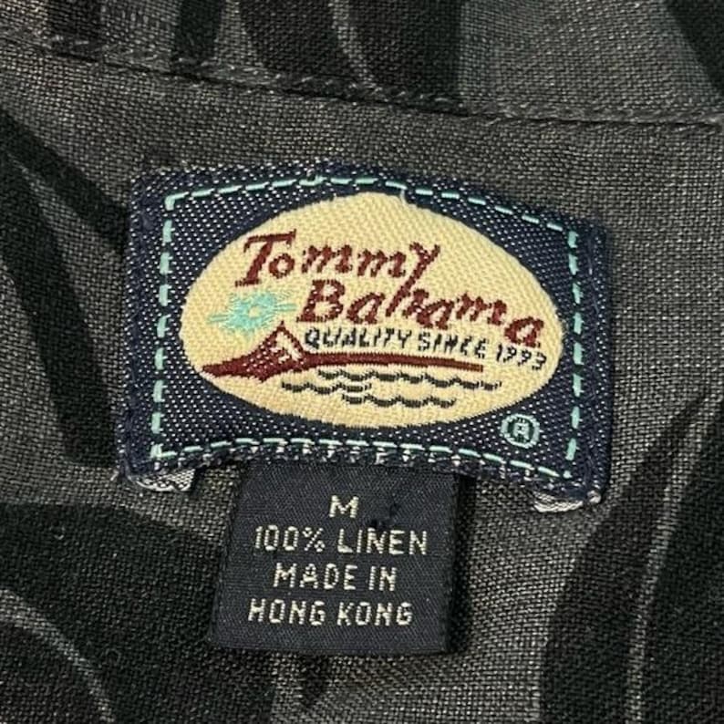 Puede incluir: Primer plano de una camisa gris oscuro con un estampado floral. La etiqueta Tommy Bahama es visible, con el nombre de la marca, un velero y el texto "QUALITY SINCE 1993". La etiqueta indica que la camisa es 100% lino y est&aacute; hecha en Hong Kong.