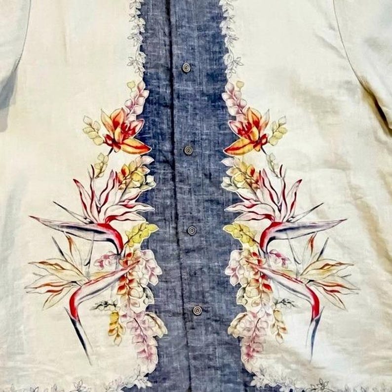 May include: A button-down shirt with a cream-colored background and a central vertical panel of blue fabric. The shirt features a floral pattern with red, yellow, and white flowers and bird of paradise designs. The shirt has a classic collar and short sleeves.