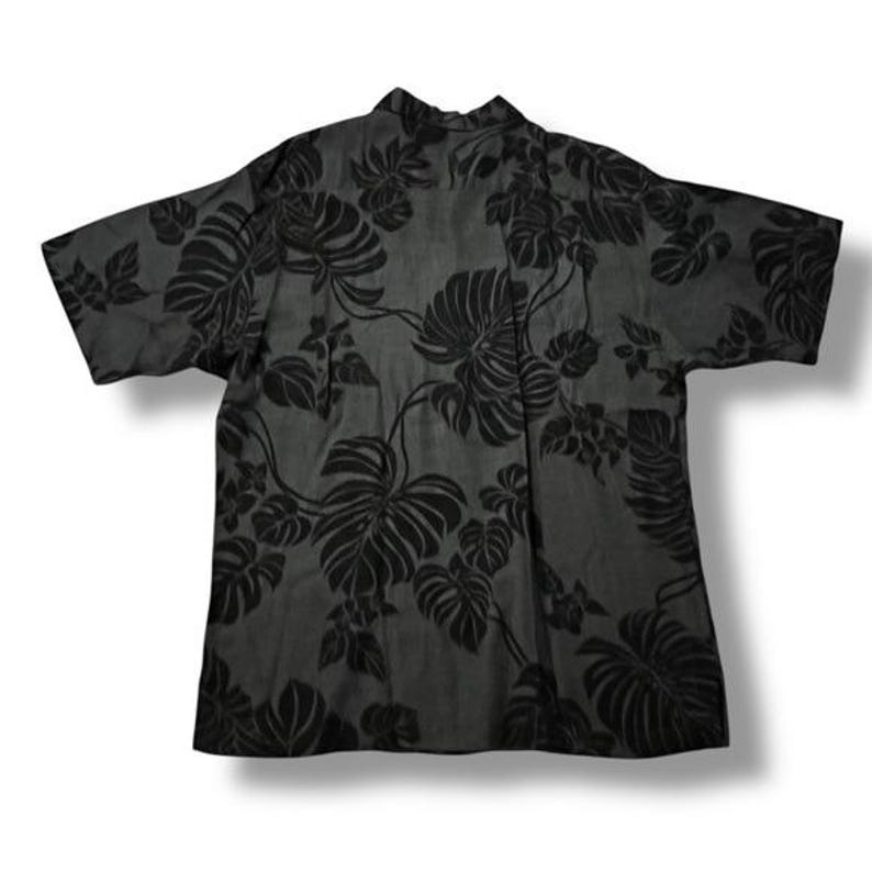 Puede incluir: Una camisa gris oscuro con un estampado de hojas tropicales negras. La camisa de manga corta presenta un cuello cl&aacute;sico y un corte relajado, adecuada para ropa informal. La camisa est&aacute; hecha de una tela ligera.