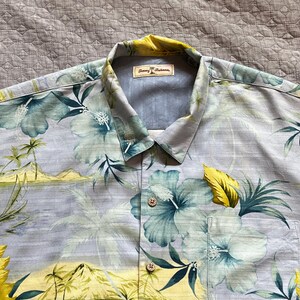 May include: A light blue button-down shirt with a tropical floral print. The shirt features large teal hibiscus flowers, yellow accents, and a landscape design. The shirt has a collar and a pocket. The label reads "Tommy Bahama."