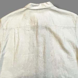 May include: Back view of a light beige button-down shirt. The shirt has a collar and a yoke detail across the upper back. The fabric appears to be linen or a similar lightweight material.