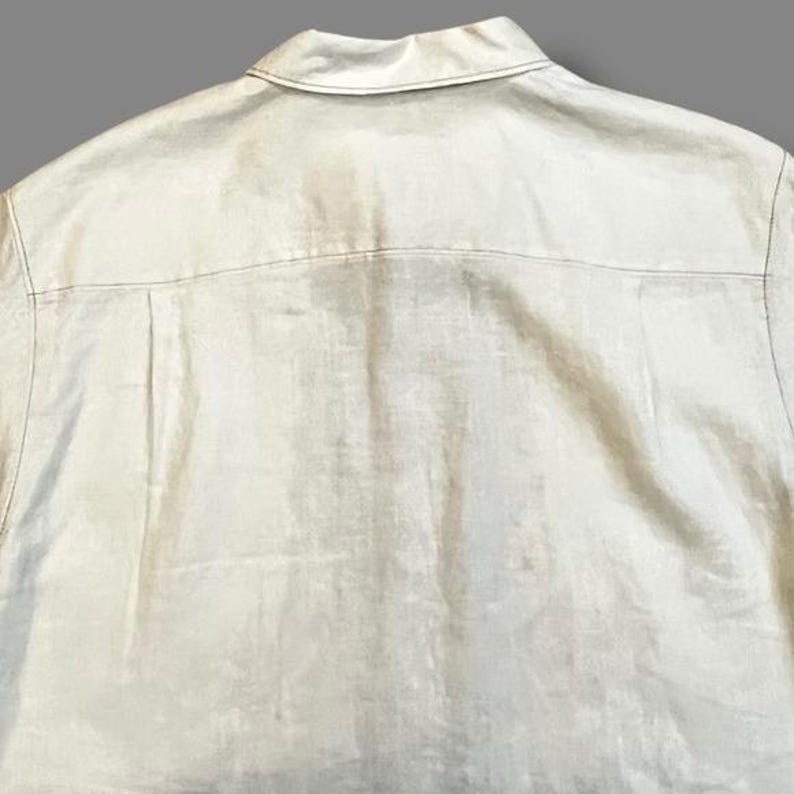 May include: Back view of a light beige button-down shirt. The shirt has a collar and a yoke detail across the upper back. The fabric appears to be linen or a similar lightweight material.