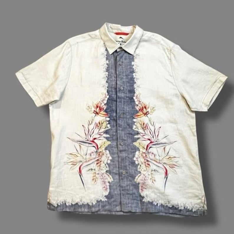 May include: Short-sleeved button-down shirt with a white body and a blue center panel. The shirt features a floral design with red, yellow, and green accents. The shirt has a collared neckline and a relaxed fit.
