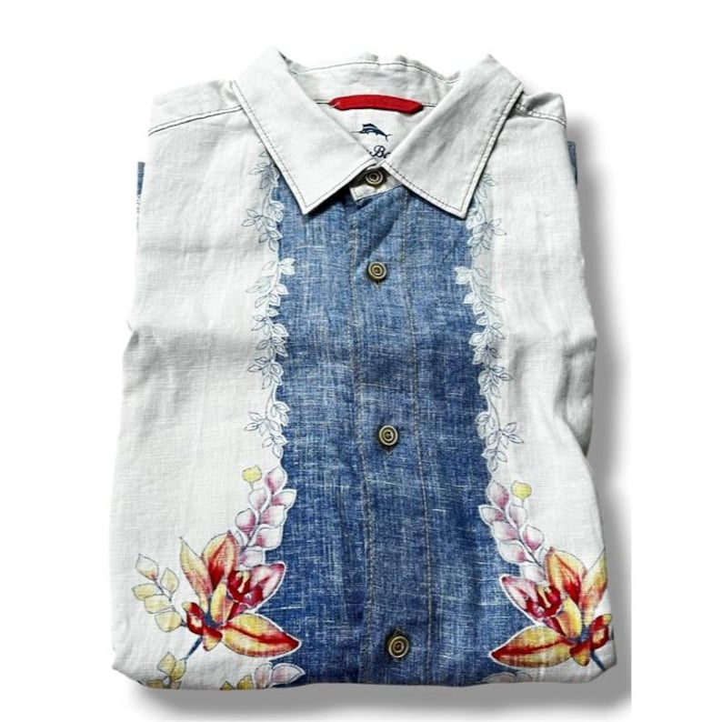 May include: A folded button-down shirt with a white collar and a blue denim-like center panel. The shirt features floral patterns in red, yellow, and pink, with a white background. The shirt has three brown buttons and a red tag near the collar.