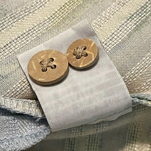 May include: Close-up of a light blue and white plaid shirt with two round, light brown coconut shell buttons. The buttons are attached to a light gray fabric tab.