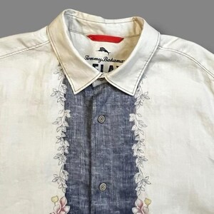 May include: Cream-colored button-up shirt with a blue center panel and floral leaf design. The shirt features the Tommy Bahama logo on the collar. Two buttons are visible on the front. The shirt is made of a lightweight fabric.