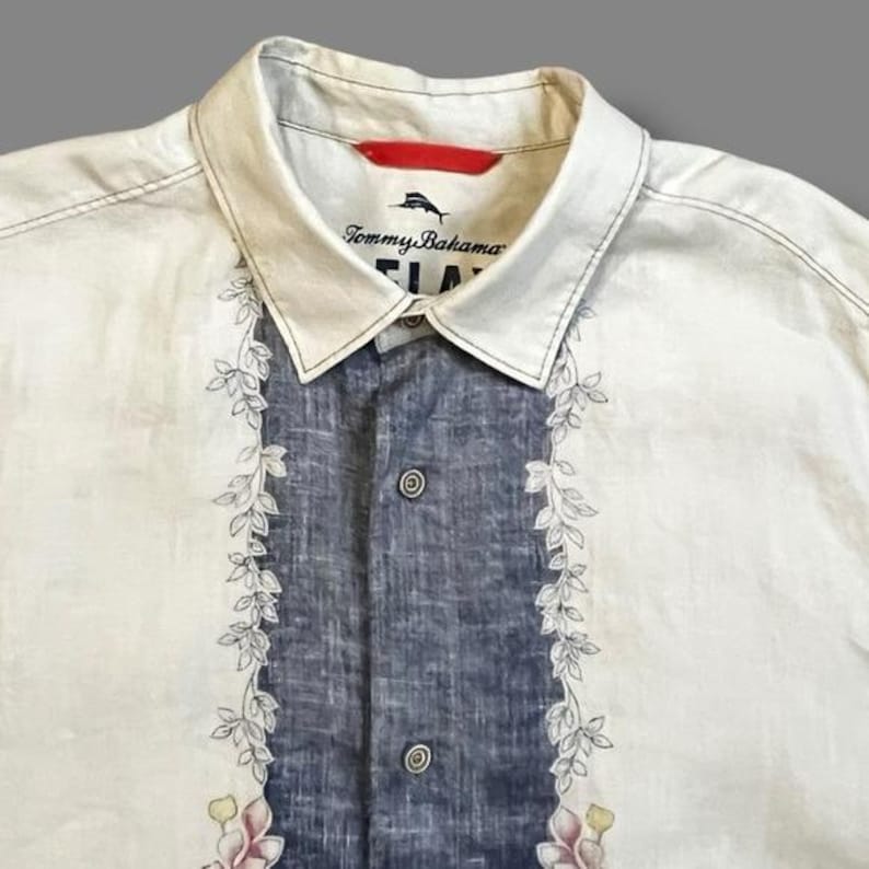 May include: Cream-colored button-up shirt with a blue center panel and floral leaf design. The shirt features the Tommy Bahama logo on the collar. Two buttons are visible on the front. The shirt is made of a lightweight fabric.