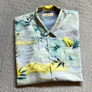May include: A light blue button-down shirt with a tropical floral print. The shirt features a pattern of large teal and yellow hibiscus flowers, palm trees, and a coastal landscape. The collar and pocket are visible.