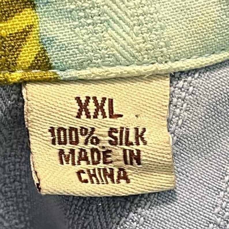 May include: Close-up of a fabric tag with the text "XXL", "100% SILK", and "MADE IN CHINA". The tag is beige and attached to a light blue and patterned fabric.