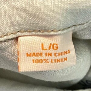 May include: Close-up of a peach-colored tag with the text "L/G", "MADE IN CHINA", and "100% LINEN". The tag is attached to a light-colored fabric, possibly linen.