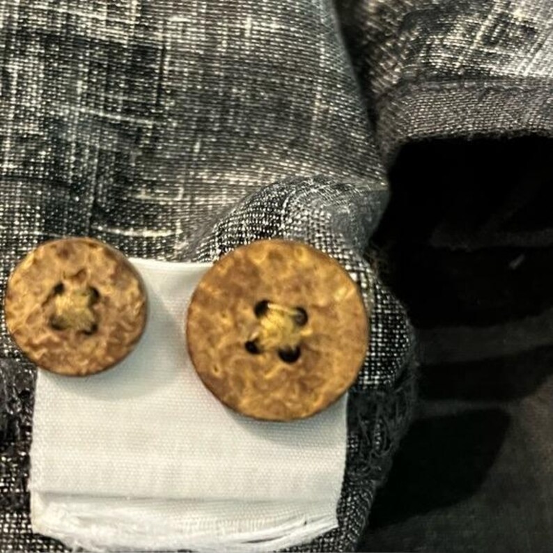 Puede incluir: Primer plano de un detalle de prenda, mostrando dos botones redondos de madera texturizada. Los botones est&aacute;n cosidos a una tela gris y blanca moteada, con una etiqueta blanca debajo. Los botones tienen un aspecto natural y r&uacute;stico.