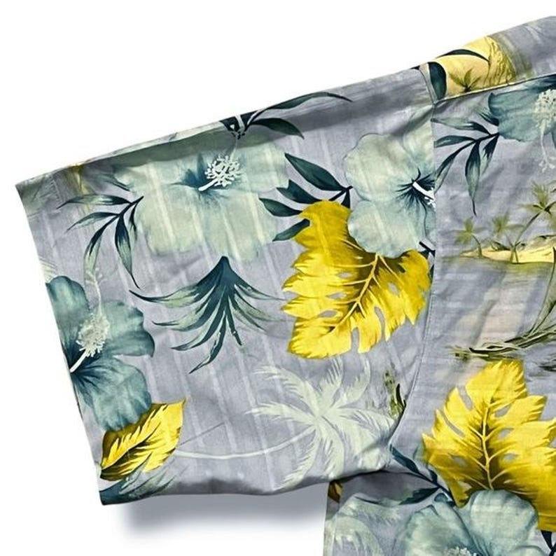 May include: A close-up of a gray button-down shirt with a tropical floral pattern. The design features large yellow and teal leaves and flowers, with palm trees and a beach scene in the background. The shirt's fabric has a subtle striped texture.