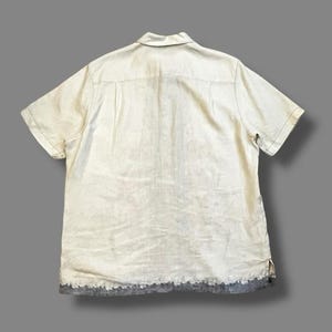 May include: A short-sleeved, off-white button-down shirt with a collar. The shirt has a faded appearance and a distressed, gray hemline. The fabric appears to be linen or a similar lightweight material. The shirt is laid flat.
