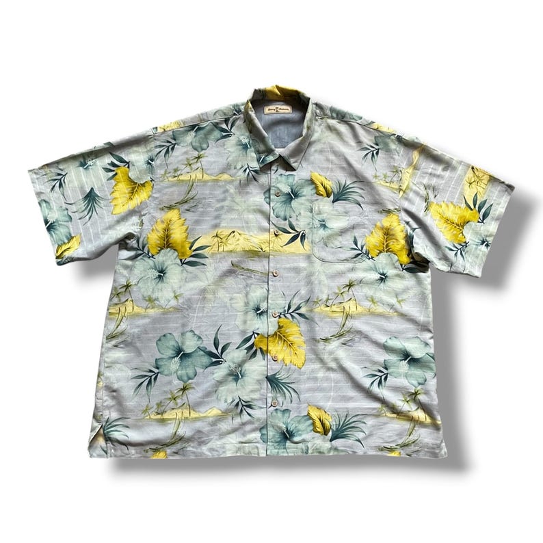 May include: A short-sleeved button-down shirt with a floral pattern. The shirt features a light blue background with yellow and teal floral and leaf designs. The shirt has a collared neckline and a single chest pocket.