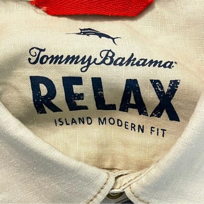 May include: Close-up of a cream-colored shirt collar with the Tommy Bahama logo and the word "RELAX" in navy blue block letters. The text "ISLAND MODERN FIT" is below the word "RELAX". A red tag is visible at the top.