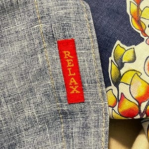 May include: Close-up of a blue and white textured fabric with a red rectangular tag that reads "RELAX" in white lettering. Part of a floral patterned fabric is visible on the right side.