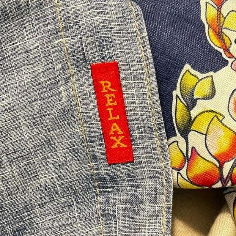May include: Close-up of a blue and white textured fabric with a red rectangular tag that reads "RELAX" in white lettering. Part of a floral patterned fabric is visible on the right side.