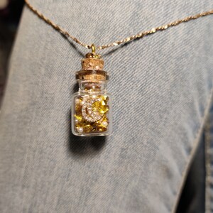Op de afbeelding: Een gouden ketting met een klein glazen flesje hanger gevuld met gouden en witte glitter en een gouden halve maan bedel.