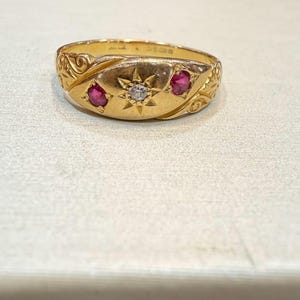 Peut inclure: Une bague en or avec un motif central en forme d'éclat d'étoile, un petit diamant et deux pierres précieuses rouges. La bague présente des motifs tourbillonnants et une finition polie. Un bijou de style vintage.