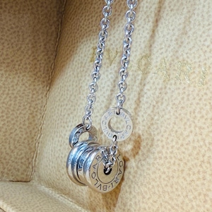 May include: A silver-toned necklace featuring a chain and a pendant with multiple stacked, cylindrical elements. The pendant has engraved lettering. The necklace is displayed against a neutral background.