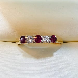 May include: A gold ring with five gemstones. It alternates between round, deep red rubies and brilliant white diamonds. The ring is set against a soft, off-white background, accentuating the contrast of the gemstones.