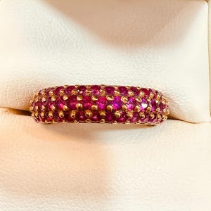 Op de afbeelding: Een gouden ring met een band bedekt met kleine, ronde, dieproze edelstenen. De ring staat tegen een witte achtergrond, waardoor de levendige kleur van de edelstenen en de gouden zetting worden benadrukt.